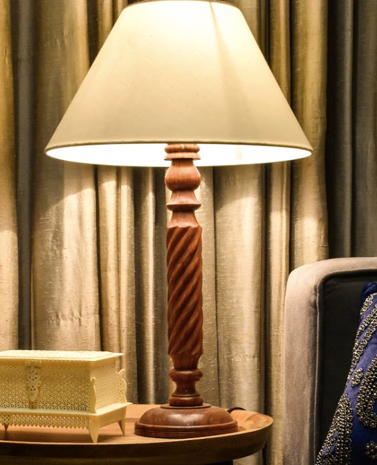 A wooden lamp made with mango wood, carved with elegant detailing along with a beige lamp shade.