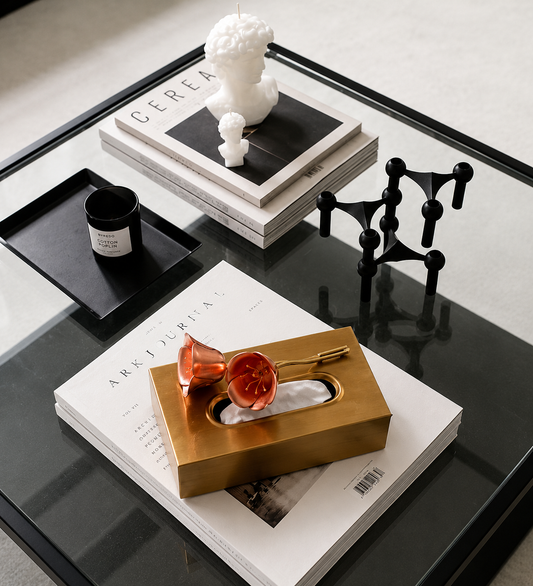 A striking rectangular tissue box in a brushed brass-plated finish, topped with an oversized resin flower in a warm coral-red hue that curls delicately across the lid. 