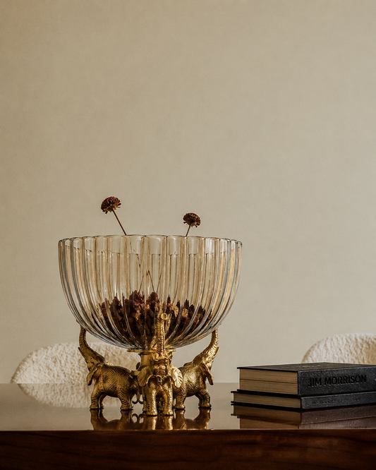 A statement decorative bowl where three brass elephants stand trunk-to-tusk, their raised trunks cradling a fluted, amber-tinted glass bowl above.