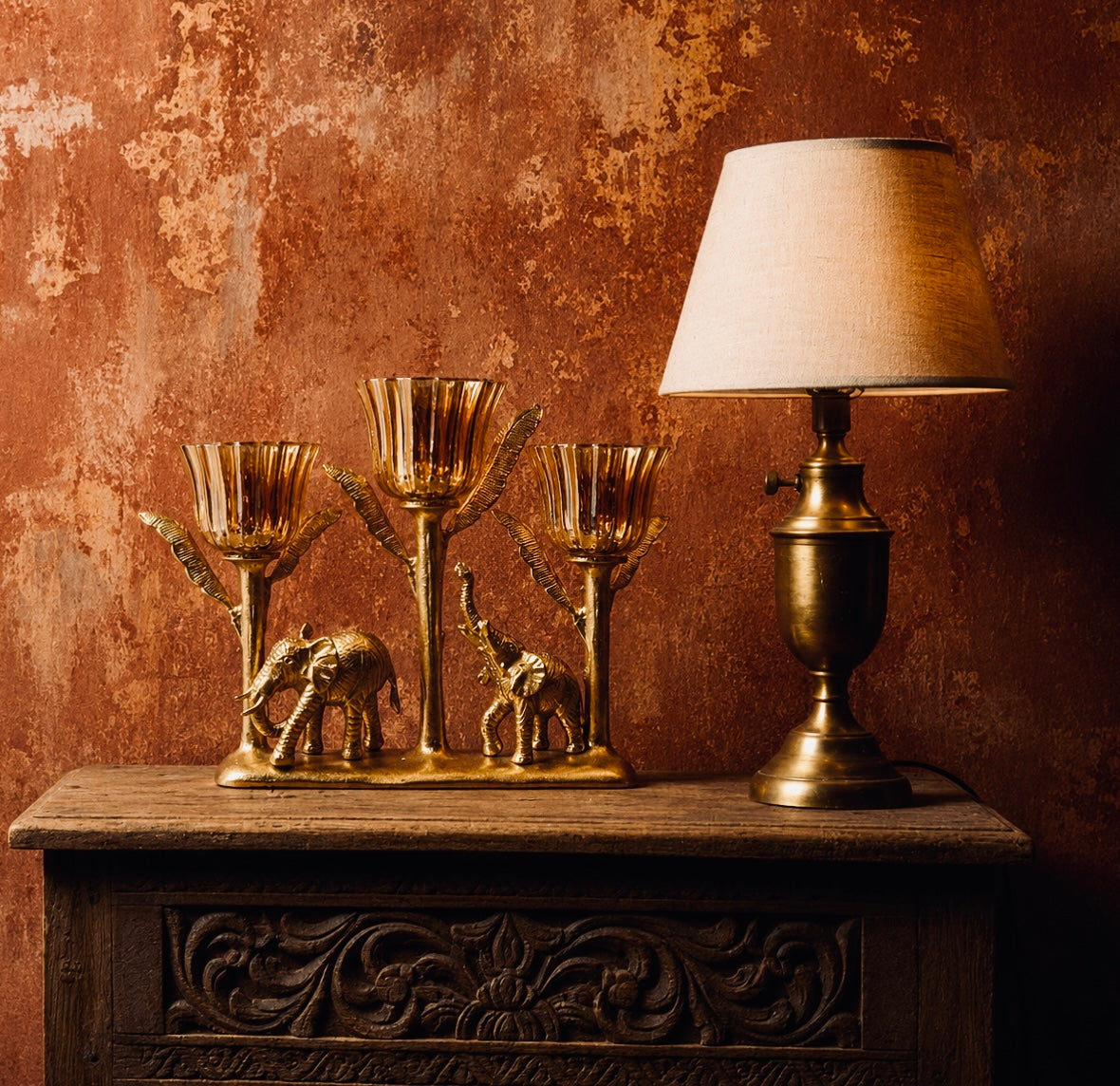 A striking three-arm candelabra where a mother and calf elephant walk beneath a canopy of leaf-shaped brass stems, each topped with a fluted amber glass votive cup. 