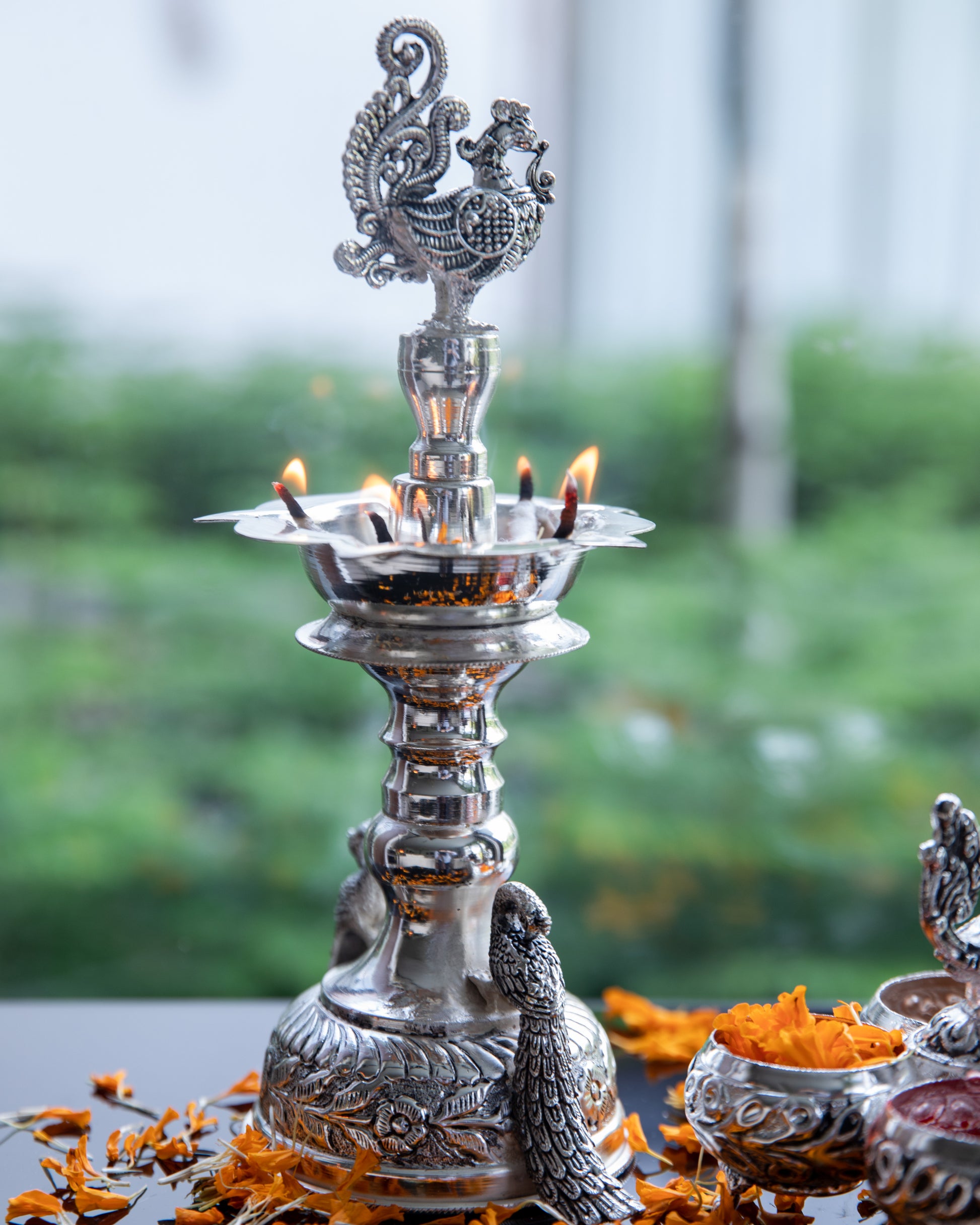 Our Silver Peacock Tall Diya is coated with premium silver plating, this diya stand radiates a lustrous and timeless glow when the diya is lit. It holds five wicks of diya.
