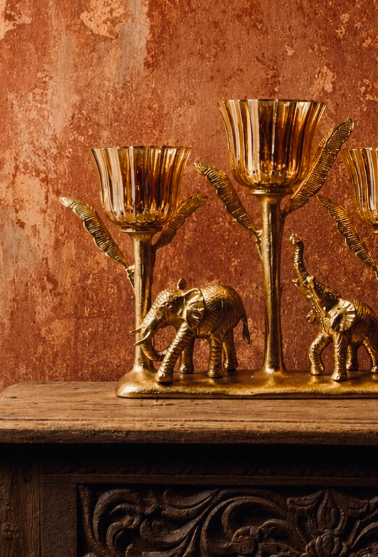A striking three-arm candelabra where a mother and calf elephant walk beneath a canopy of leaf-shaped brass stems, each topped with a fluted amber glass votive cup. 