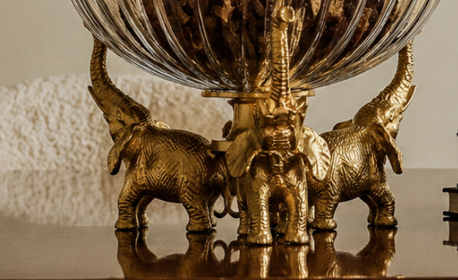 A statement decorative bowl where three brass elephants stand trunk-to-tusk, their raised trunks cradling a fluted, amber-tinted glass bowl above.