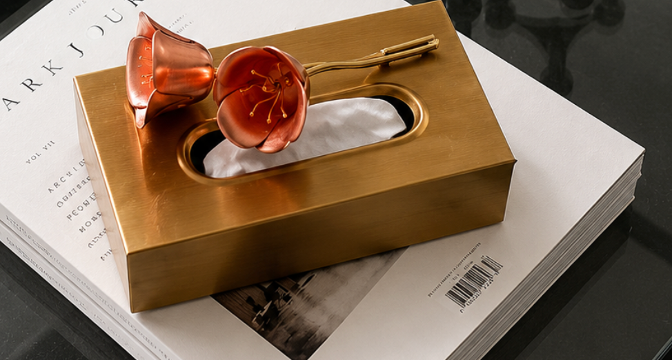 A striking rectangular tissue box in a brushed brass-plated finish, topped with an oversized resin flower in a warm coral-red hue that curls delicately across the lid. 