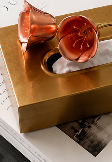 A striking rectangular tissue box in a brushed brass-plated finish, topped with an oversized resin flower in a warm coral-red hue that curls delicately across the lid. 